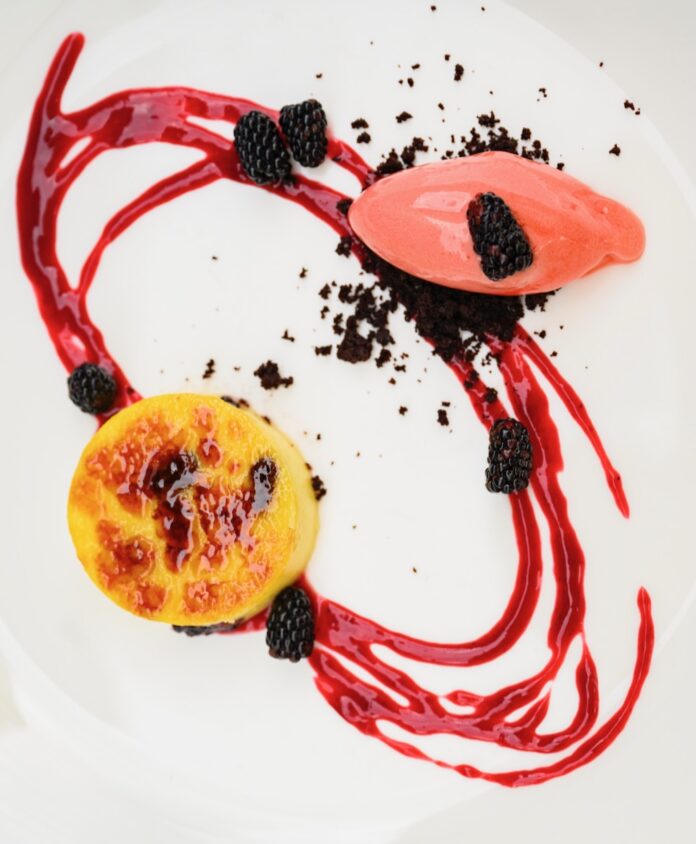 A white plate holds a blackberry creme brulee with a blackberry swirl around the plate and a small scoop of ice cream.