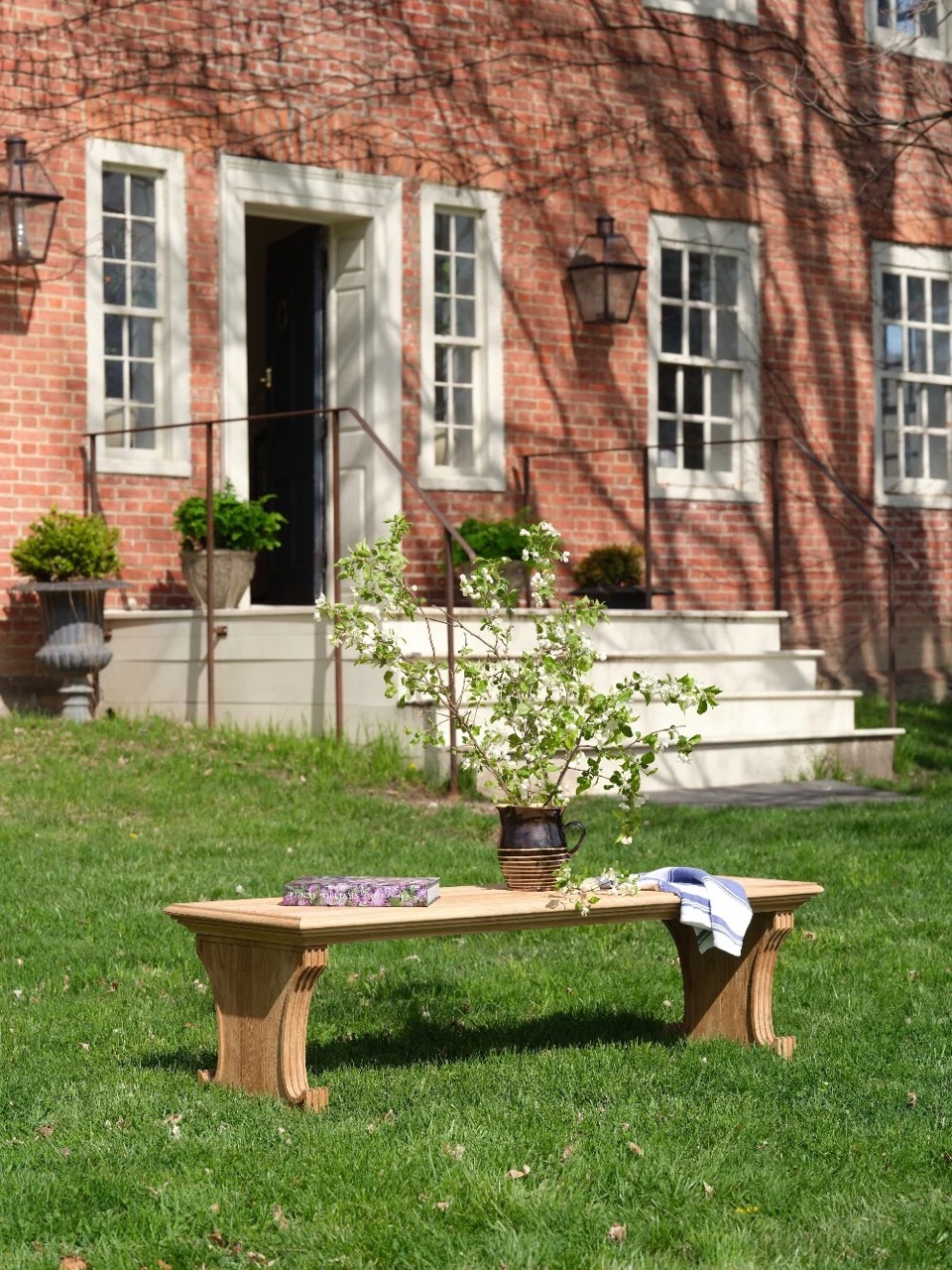A Bunny Williams brown bench sits outside with a plant, books, and a scarf sitting on it.