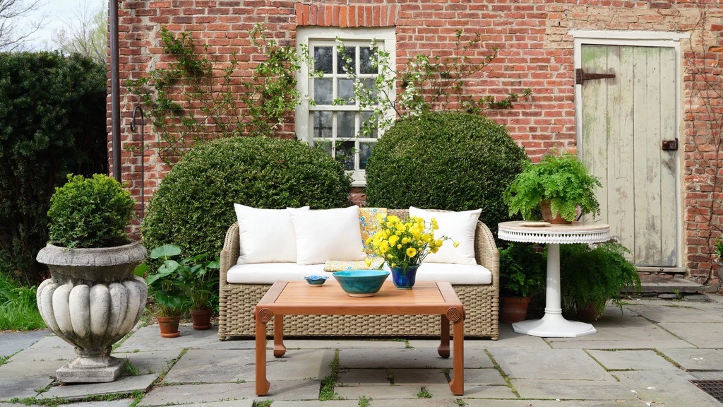 A Bunny Williams white outdoor couch sits outside on a deck behind a brown outdoor table.