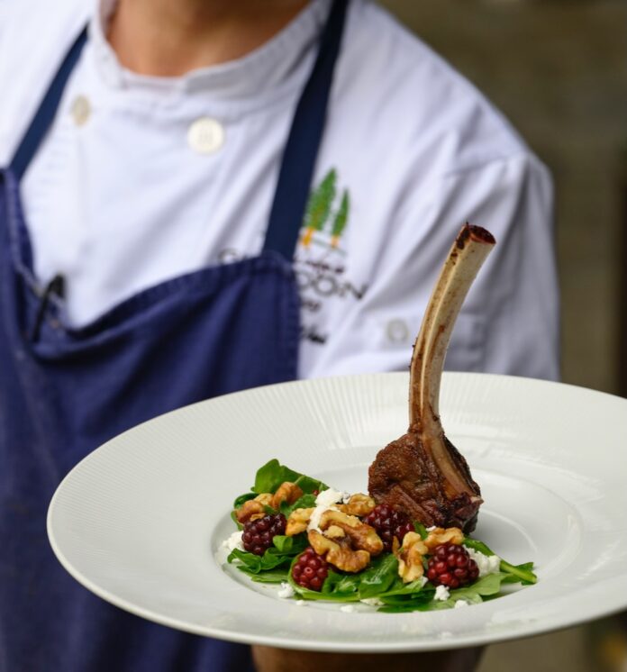 The Lodge at Glendorn Showcases Foraging to Fine Dining A man in an apron holds a leg of lamb on a white plate.