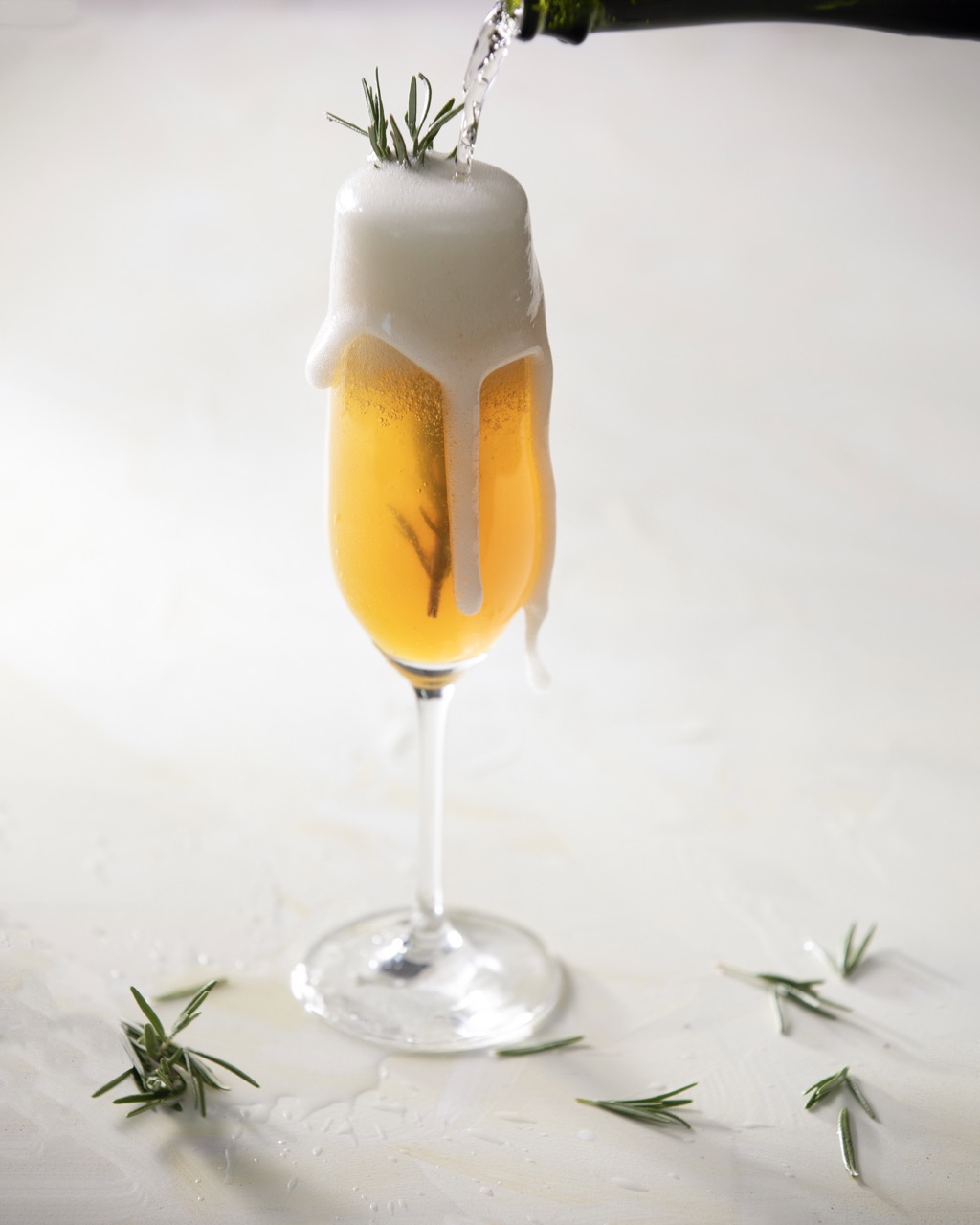 Champagne overflows into a glass with golden colored liquid and a sprig of rosemary.