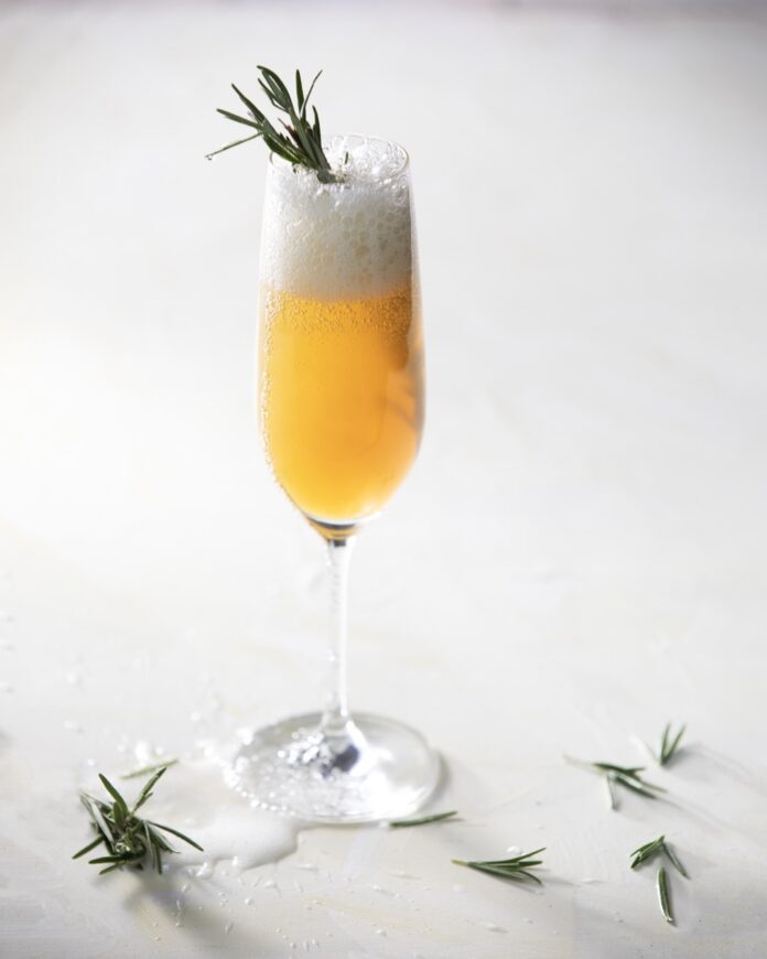 Gold Medal Cocktail A cocktail glass sits on a white background with a fizzy Gold Medal drink inside and a sprig of rosemary on top.