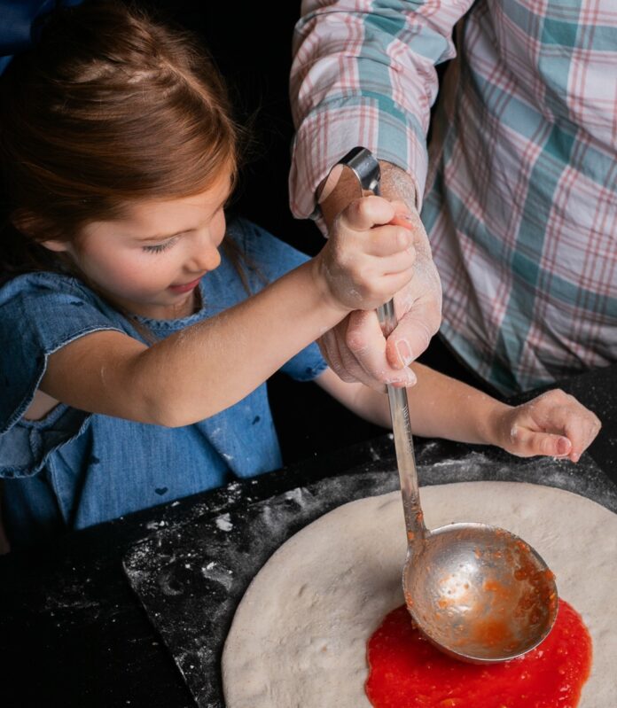 A little girl spread a simple pizza sauce onto a pizza crust with a laddle.
