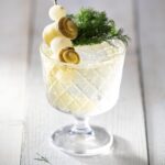 A short glass holds a pickle spritz on a white picnic table topped with rolled pickles, cocktail onions, and fresh dill.