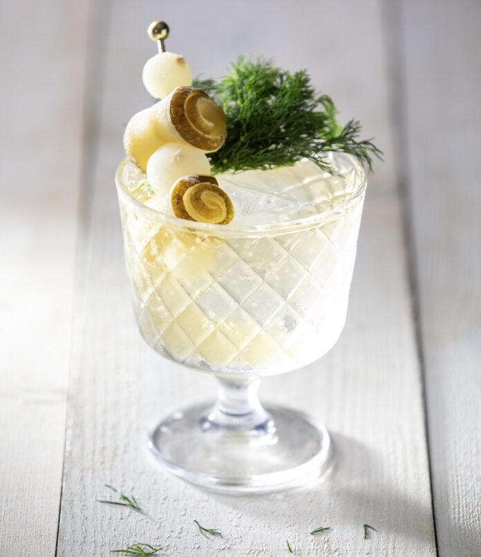 Pickle Spritz Cocktail Recipe A short glass holds a pickle spritz on a white picnic table topped with rolled pickles, cocktail onions, and fresh dill.