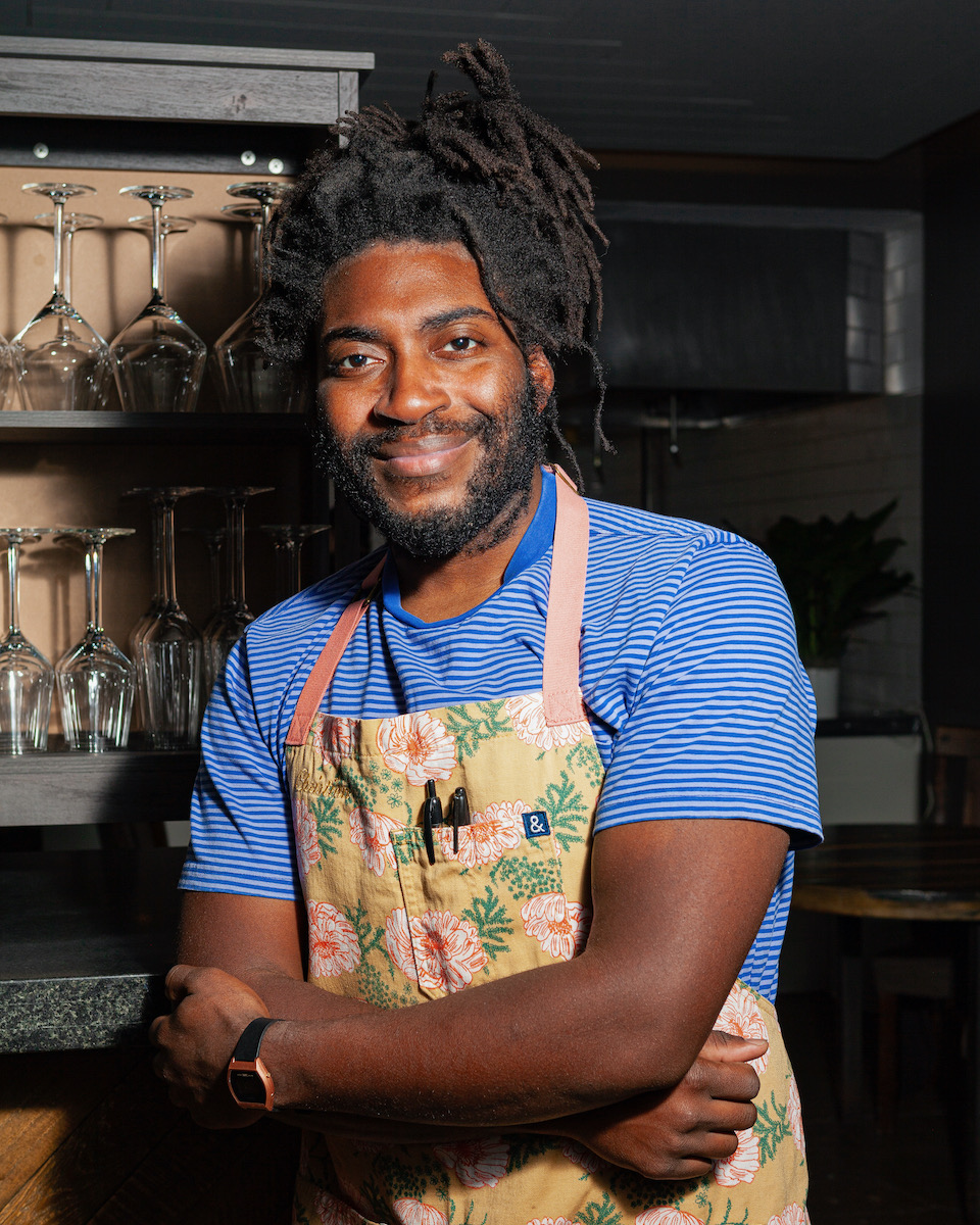 Chef Christian Hunter of Atelier, a black man with dreads in a yellow apron, stands with crossed arms