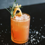 The Beso Beso, a bright pink cocktail with grapefruit and rosemary on a black table top with salt sprinkles
