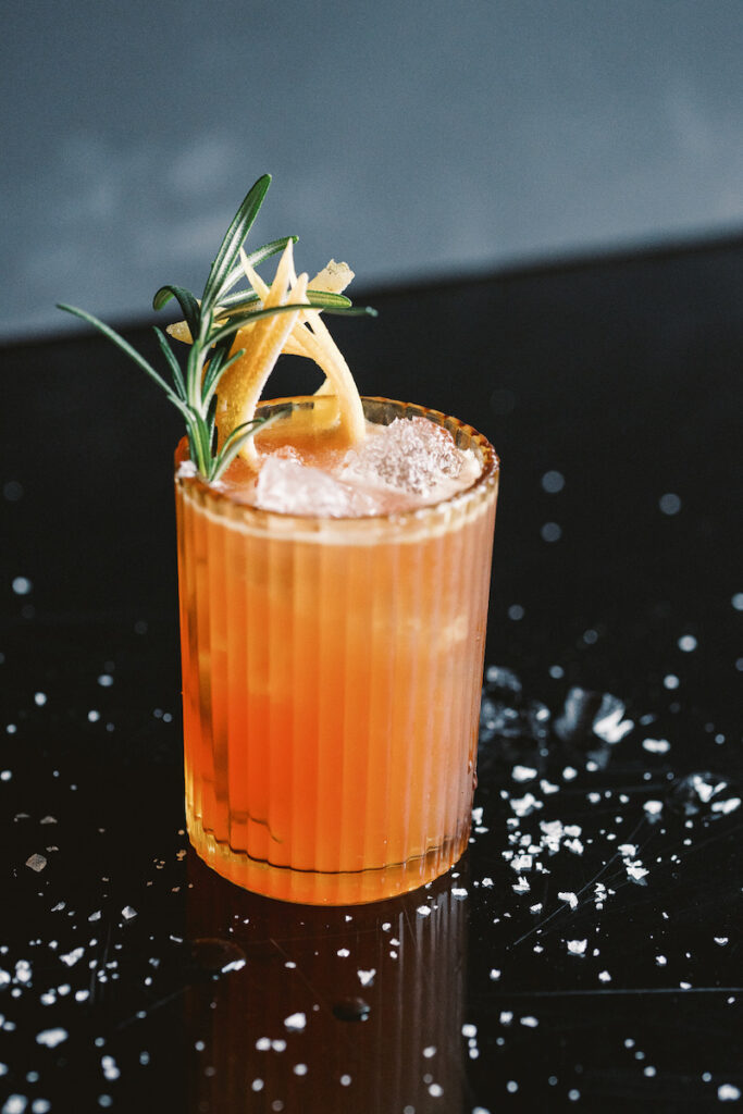 The Beso Beso, a bright pink cocktail with grapefruit and rosemary on a black table top with salt sprinkles