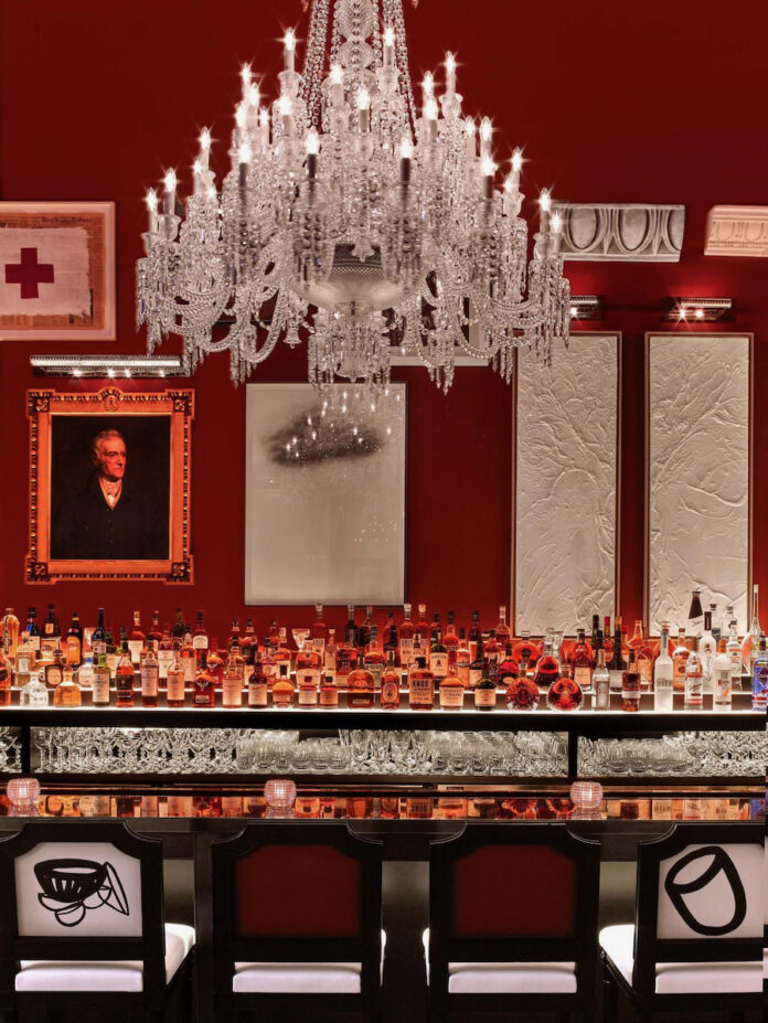A red wall with a chandelier above three bar seats at a bar in New York
