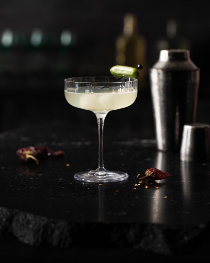 A pale Aemond House of the Dragon cocktail sits on a black table with dried chilis around the glass and a snacking cucumber as a garnish.