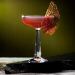A cocktail glass holds an Alien: Romulus cocktail in a red hue with a piece of tuile garnish on the outside of the glass.