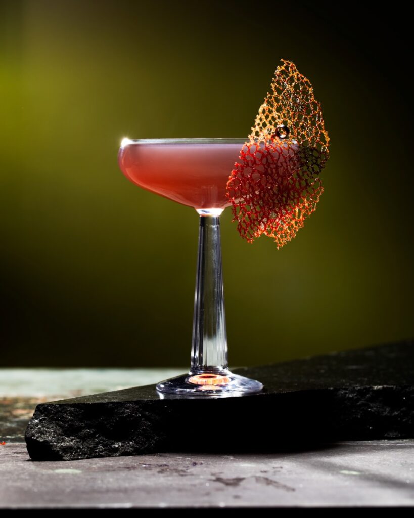 A cocktail glass holds an Alien: Romulus cocktail in a red hue with a piece of tuile garnish on the outside of the glass.