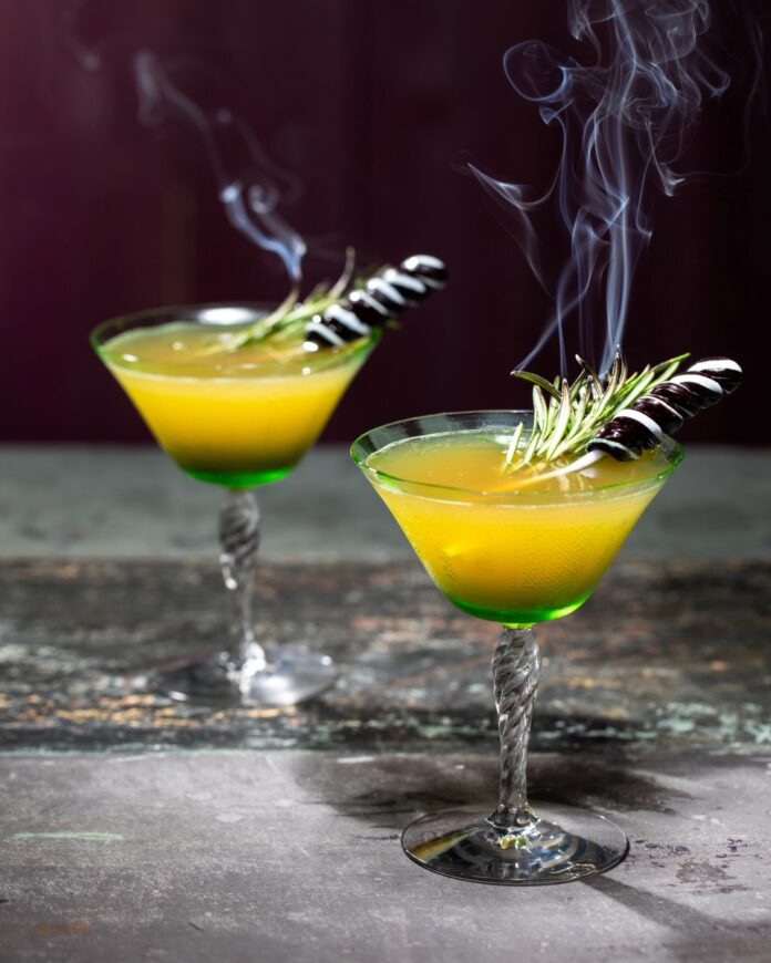 Two cocktail glasses hold a yellow cocktail with green on the bottom with a smoking rosemary and lollipop garnish on top.