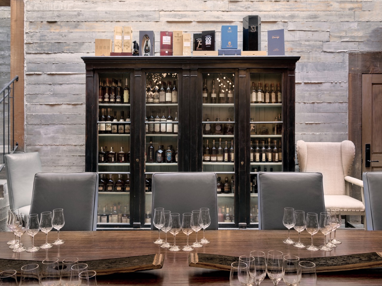 A long table sits 3 seats with 6 tasting glasses in front of them and a cabinet of wine behind them. 
