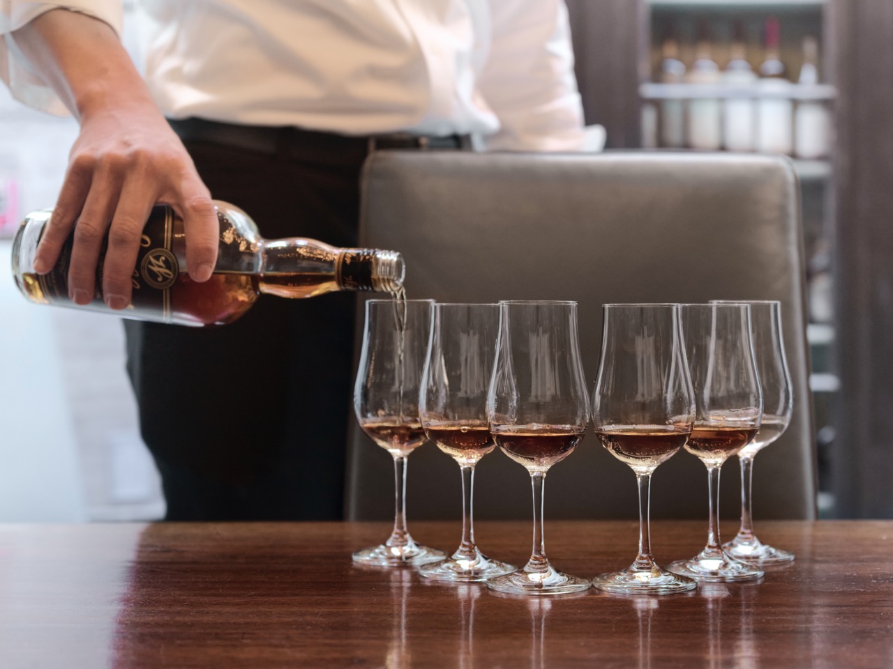 A man pours wine into a set of 6 glasses positioned in front of a chair.