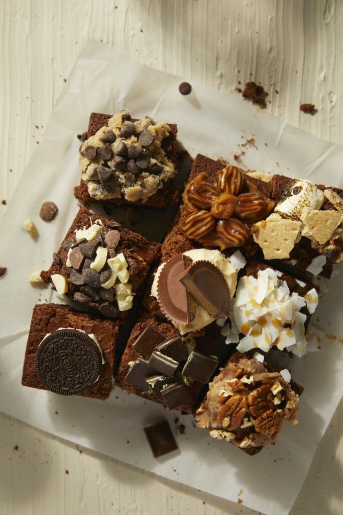 9 Boyfriend Brownies slices sit on a piece of parchment paper covered in different topping like candy, cookies, and coconut.