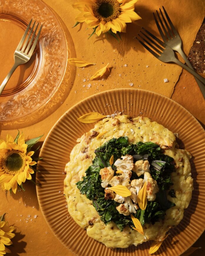 A Cauliflower Sformato sits in a bowl on an auburn table with greens on top of the sformato and cauliflower on top of that as sunflowers surround the table.
