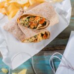 A broken in half chicken wrap sits on butcher paper with a side of chips above the broken wrap.