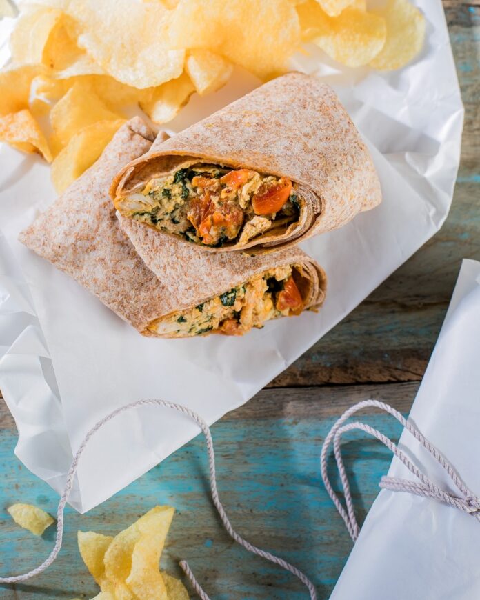 A broken in half chicken wrap sits on butcher paper with a side of chips above the broken wrap.