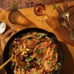 A cast iron skillet holds a creamy Chicken and Corn recipe with a spoon inside the pan and drink glasses nearby.