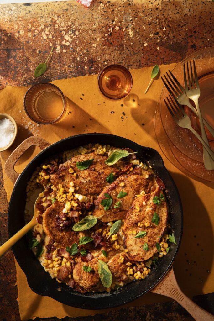 A cast iron skillet holds a creamy Chicken and Corn recipe with a spoon inside the pan and drink glasses nearby.
