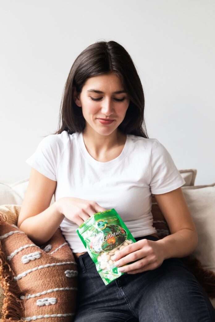 A woman sits on a couch eating a pantry snack bag of cashews.