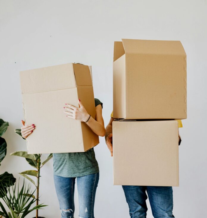 Two people carry moving boxes in front of them to move into a college dorm.