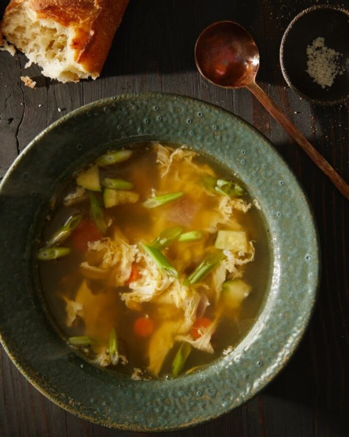 A bowl of egg drop soup with a spoon and a piece of bread on the sides