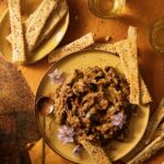 A plate holds an Eggplant Jam that sits beside an array of placed homemade crackers on another small plate.