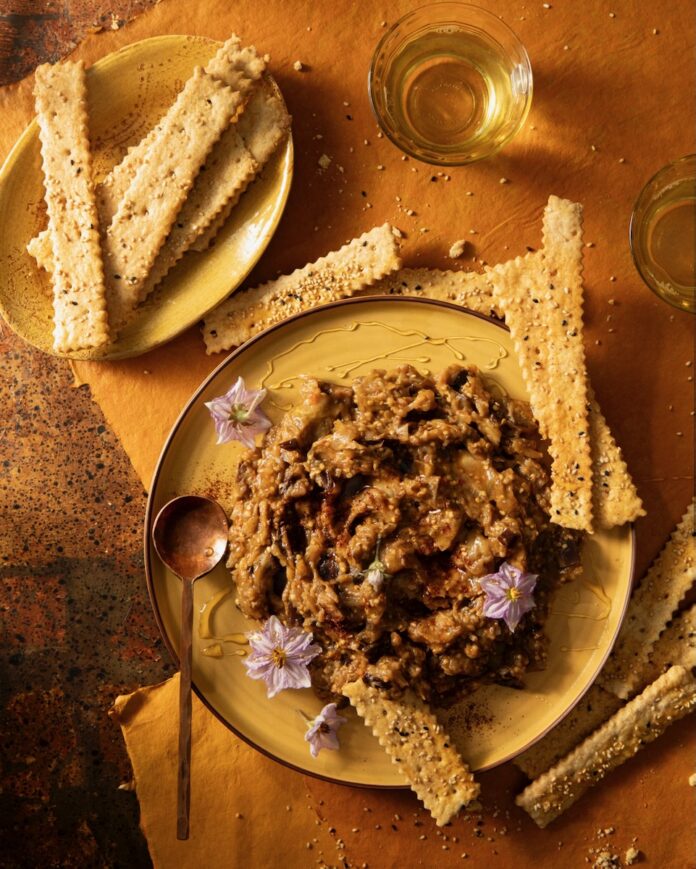 A plate holds an Eggplant Jam that sits beside an array of placed homemade crackers on another small plate.
