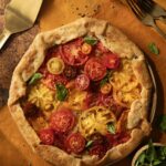 An Heirloom Tomato galette sits on a brown table with red and yellow tomatoes on top and a spatula nearby.