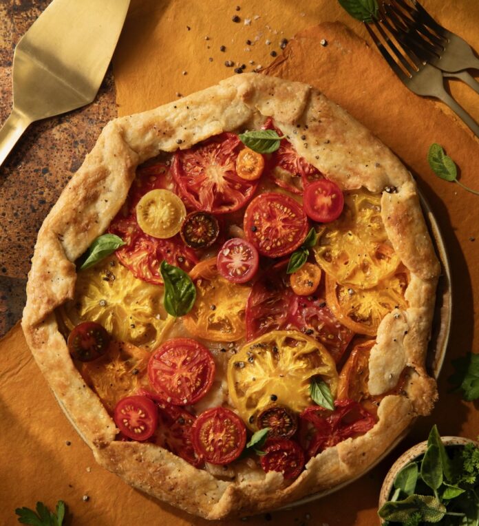 An Heirloom Tomato galette sits on a brown table with red and yellow tomatoes on top and a spatula nearby.