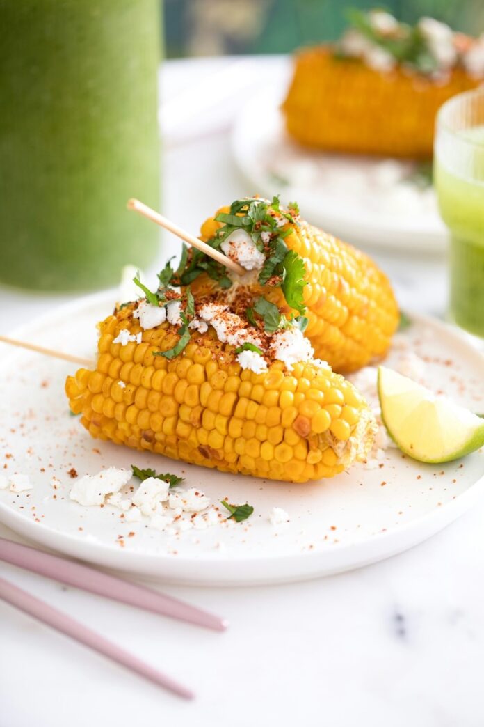 How to Cook Corn on the Cob Two corn on the cobs with cheese and herb toppings sitting beside a lime.