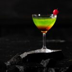 A tall cocktail glass holds a green and purple liquid with a cherry on a toothpick inside, all against a black background.
