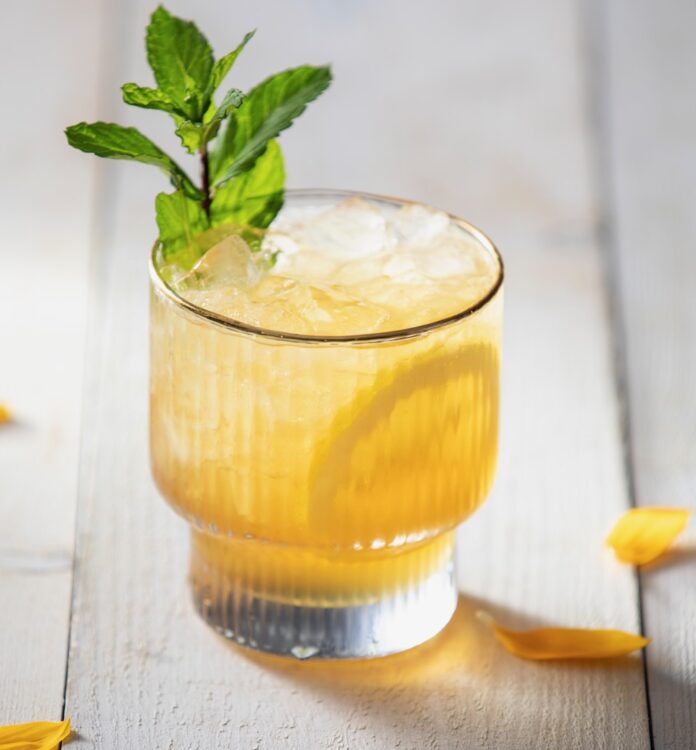 A whiskey glass holds a Meyer Lemon Smash cocktail in the color yellow topped with mint and a lemon slice all sitting on a picnic table.