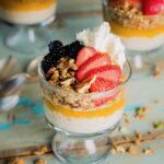 A cup of overnight oats in parfait form sits on a picnic table with oats, fresh strawberries, blackberries, and whipped cream on top.
