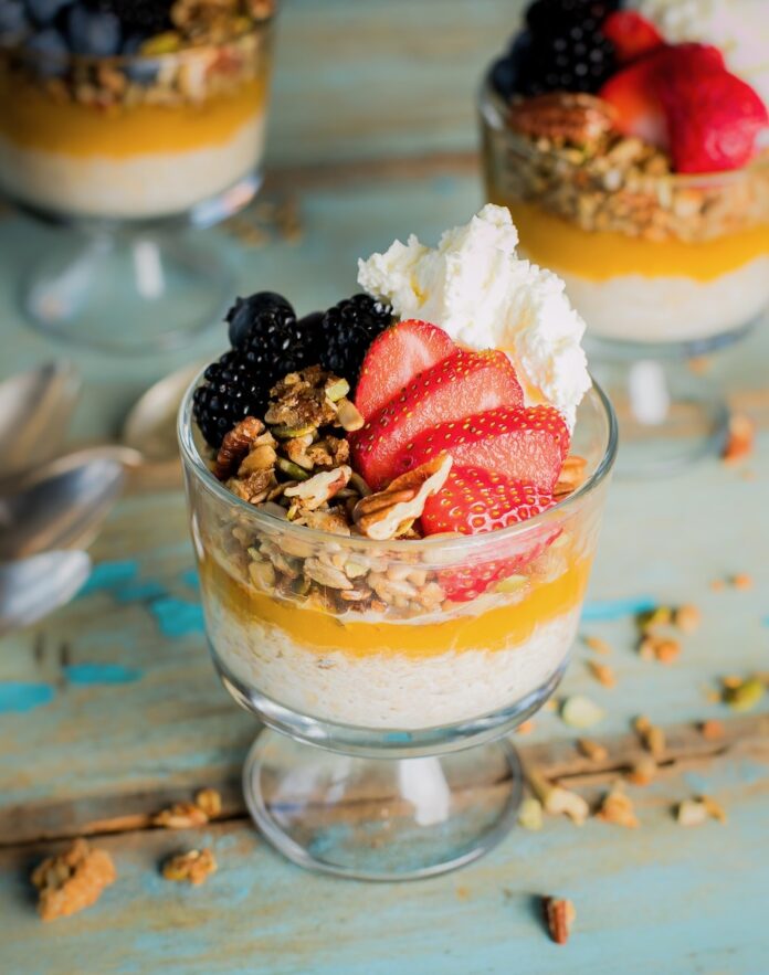 A cup of overnight oats in parfait form sits on a picnic table with oats, fresh strawberries, blackberries, and whipped cream on top.