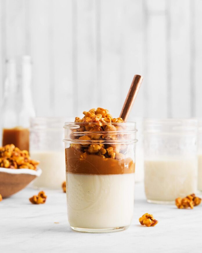 A small glass mason jar holds a popcorn panna cotta with a white creamy base, caramel, and carmael corn on top.