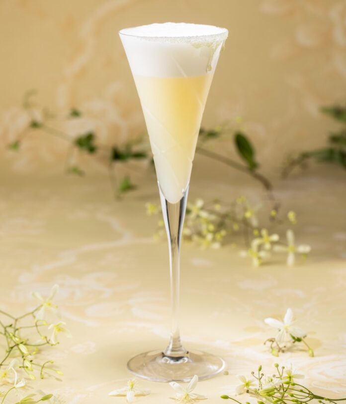 A tall glass holds a golden cocktail with foam on top representing the nenya ring from rings of power, against a floral background.