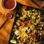 A sheet pan of roasted vegggies like corn and zucchini sits on a table next to a small bowl of spicy glaze with the spoon outside of it.