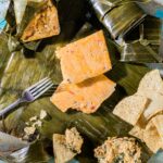 An open tamale sits on a table with the filling sitting in a square on the banana leaf accompanied by chips and wrapped tamales.