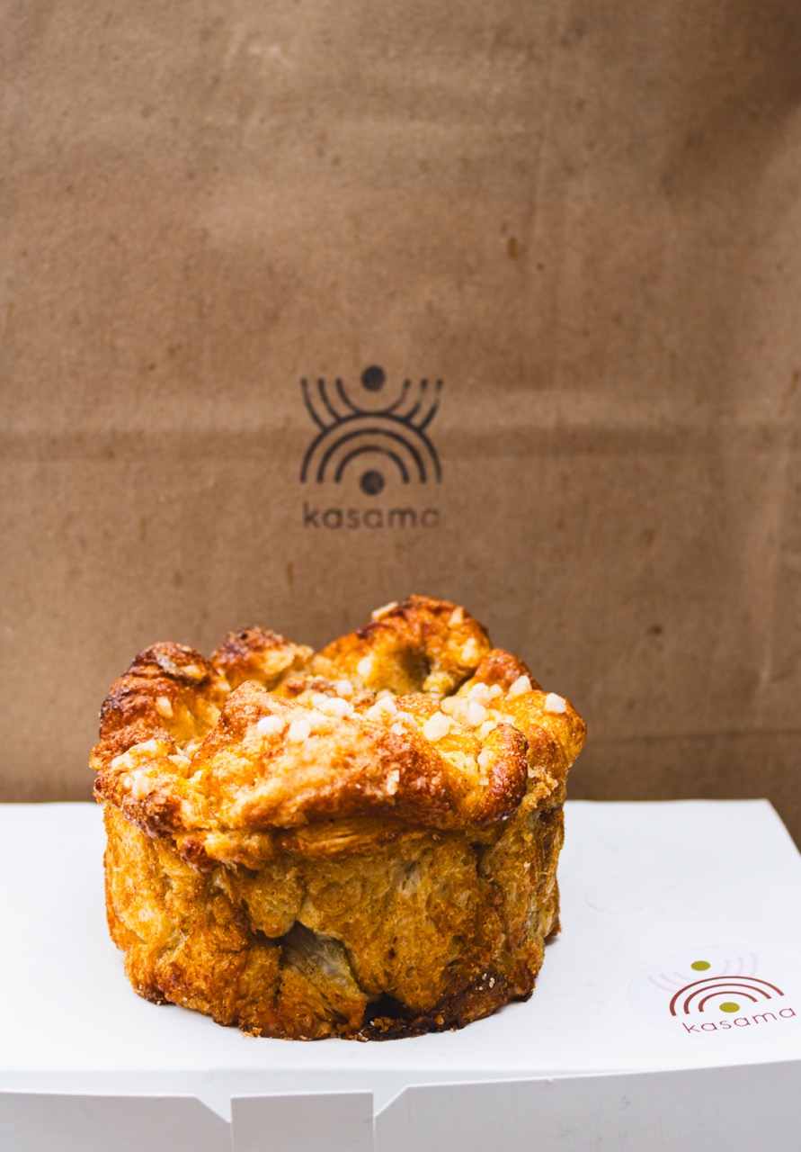 A cardamom bun from Kasama sits on top of a box in front of a brown bag with their logo on it. 