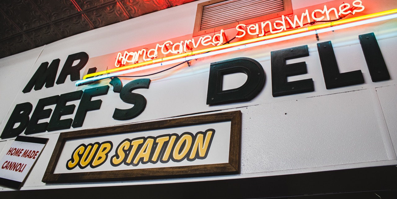 A sign for Mr. Beef's Deli sits on the wall below a red neon sign stating "handcarved sandwiches". 