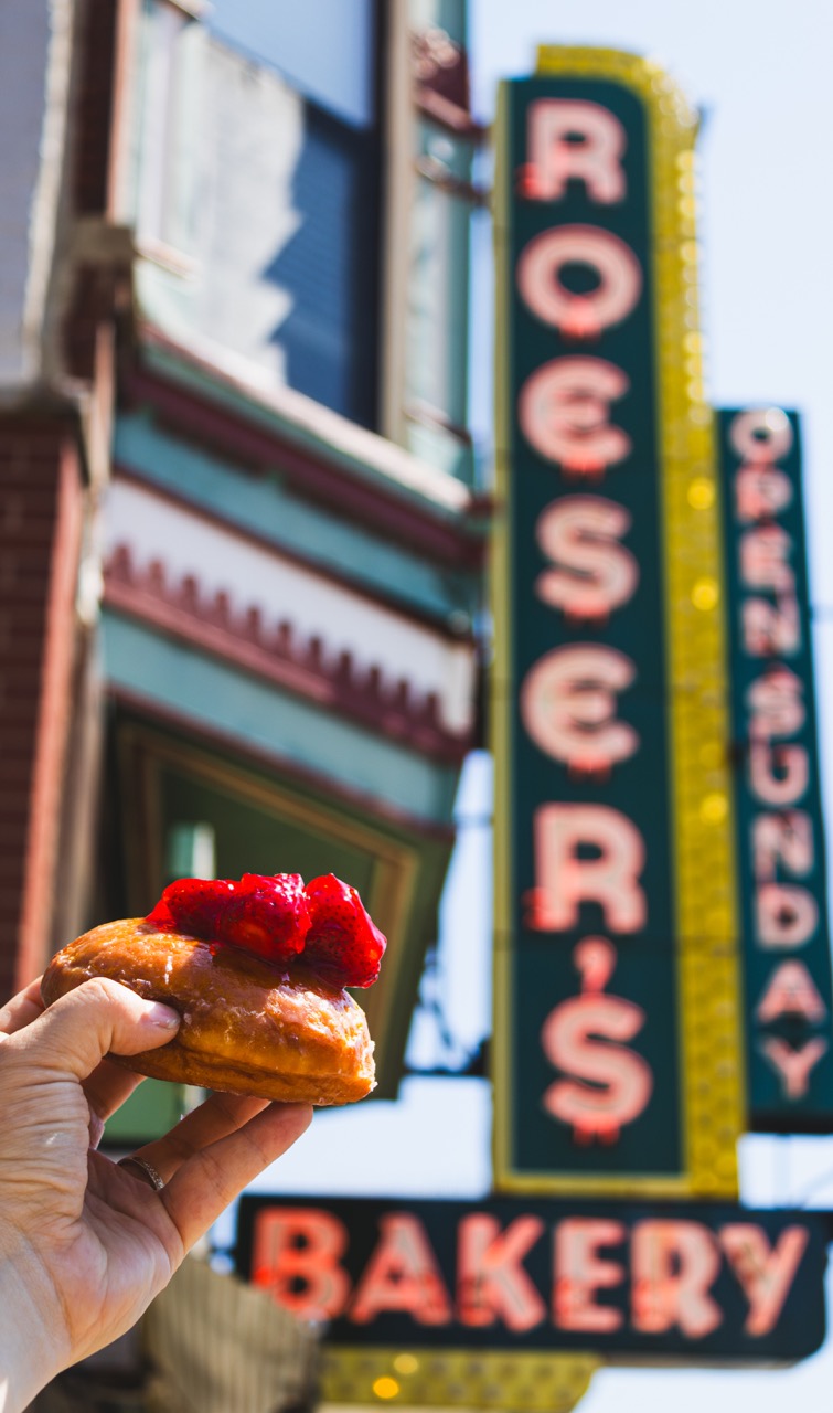 A strawberry donut featured in The Bear is held up in front of the Roeser's Bakery sign.