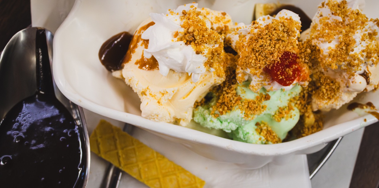 A three scoop ice cream sundae featured in The Bear sits in a white clam shell bowl next to a boat of hot fudge and a wafer cookie. 