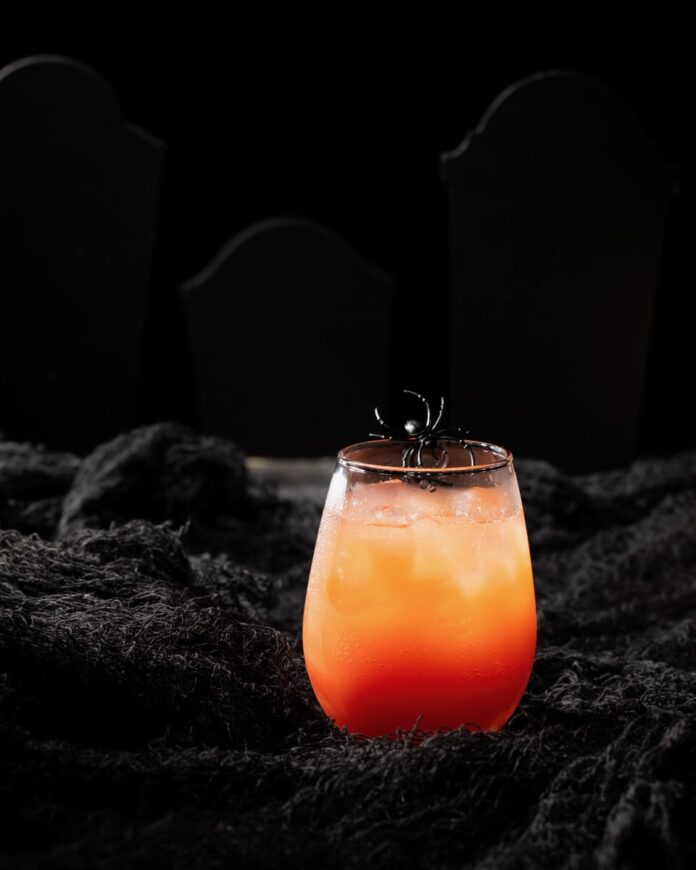 In a short cocktail glass sits a non-alcoholic spritzer in red and orange colors with a plastic spider on top.