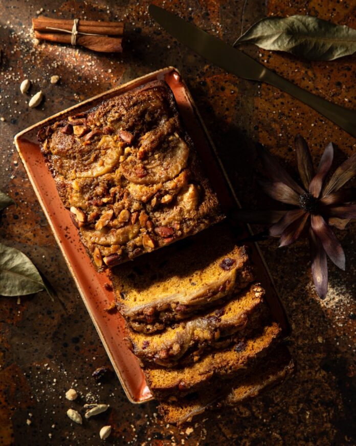 A loaf of apple cider pumpkin bread sits half sliced on a rectangular orange tray on top of a wooden table.