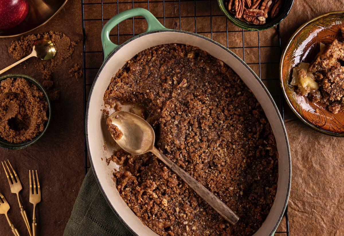 An apple crisp sits in a dish with a spoon taking a piece out of the pan. The piece sits on a plate to the right of the pan. 