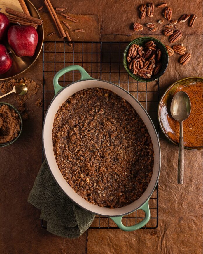 A green pan with apple crisp in it sits on a wood table surrounded by apples, brown sugar, and walnuts, and a spoon.