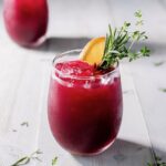 Two cocktail glasses full of beet sangria in a red color sits on a white wood table with orange slices and herb garnishes.